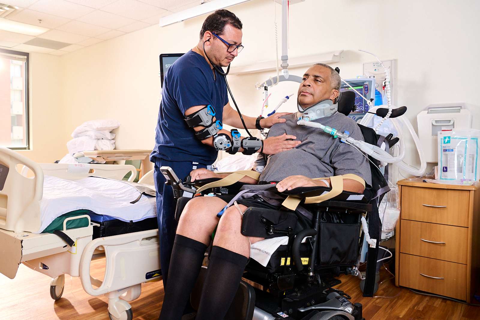 Physician uses a stethoscope to listen to a male patient's breathing. The patient is using a ventilator for breathing support following a spinal cord injury.