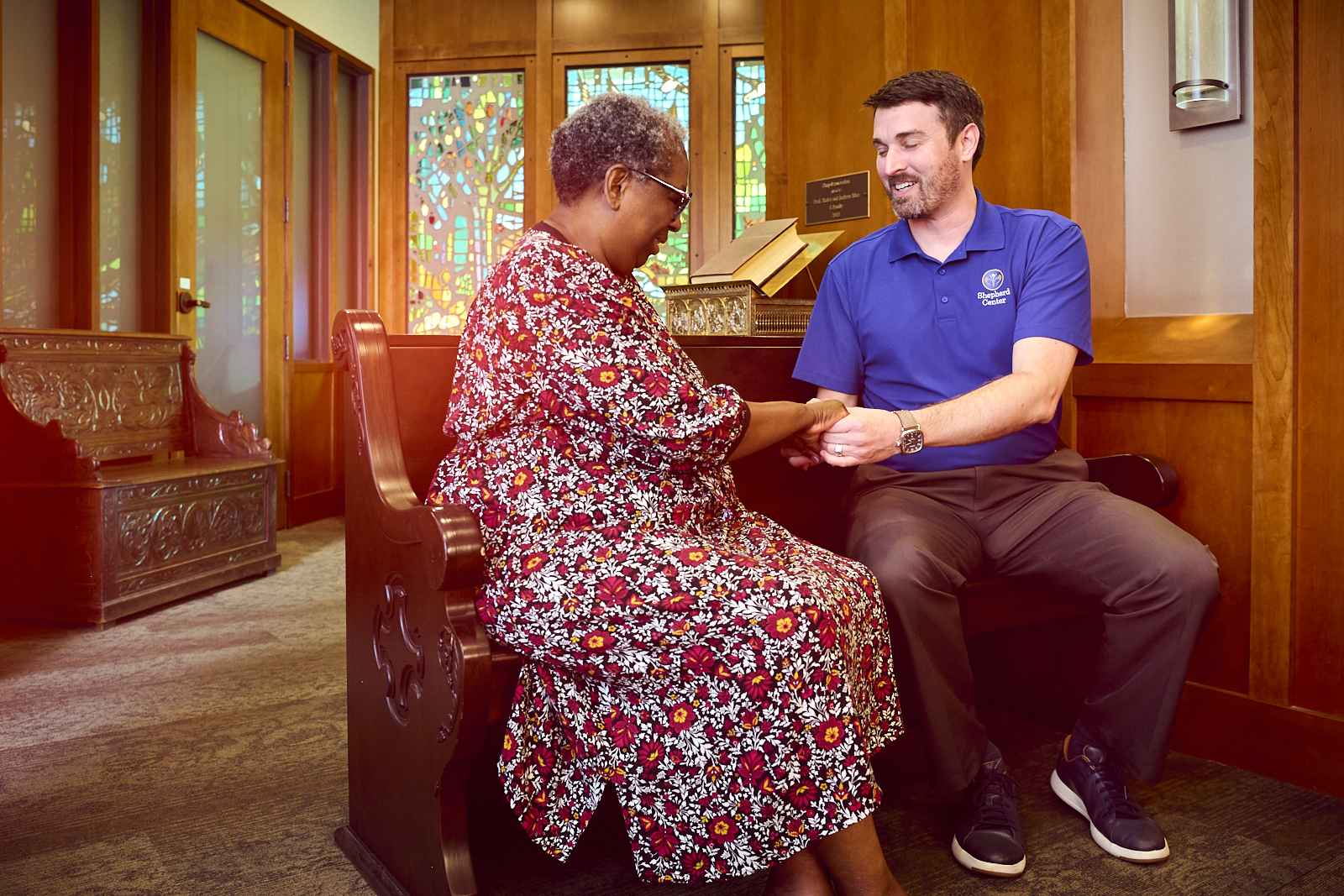 The chaplain, a man in a blue shirt is smiling while holding the hands of a woman in a floral dress who is praying. They are sitting on a wooden bench in a warmly lit chapel, with stained glass windows and wooden furnishings in the background—a serene setting.