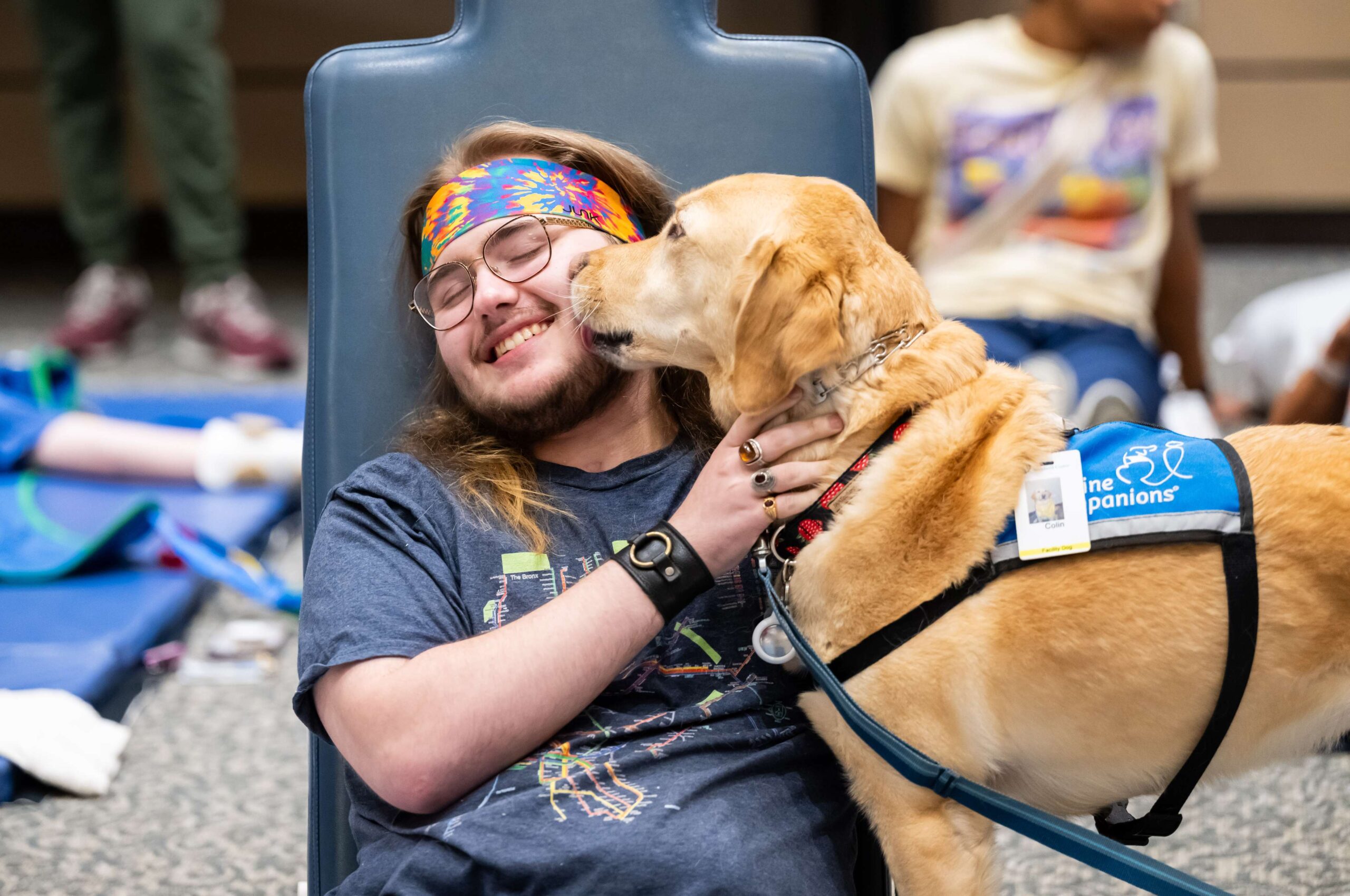 A person with long hair, glasses, and a colorful headband smiles joyfully while a yellow facility dog wearing a vest licks their face.