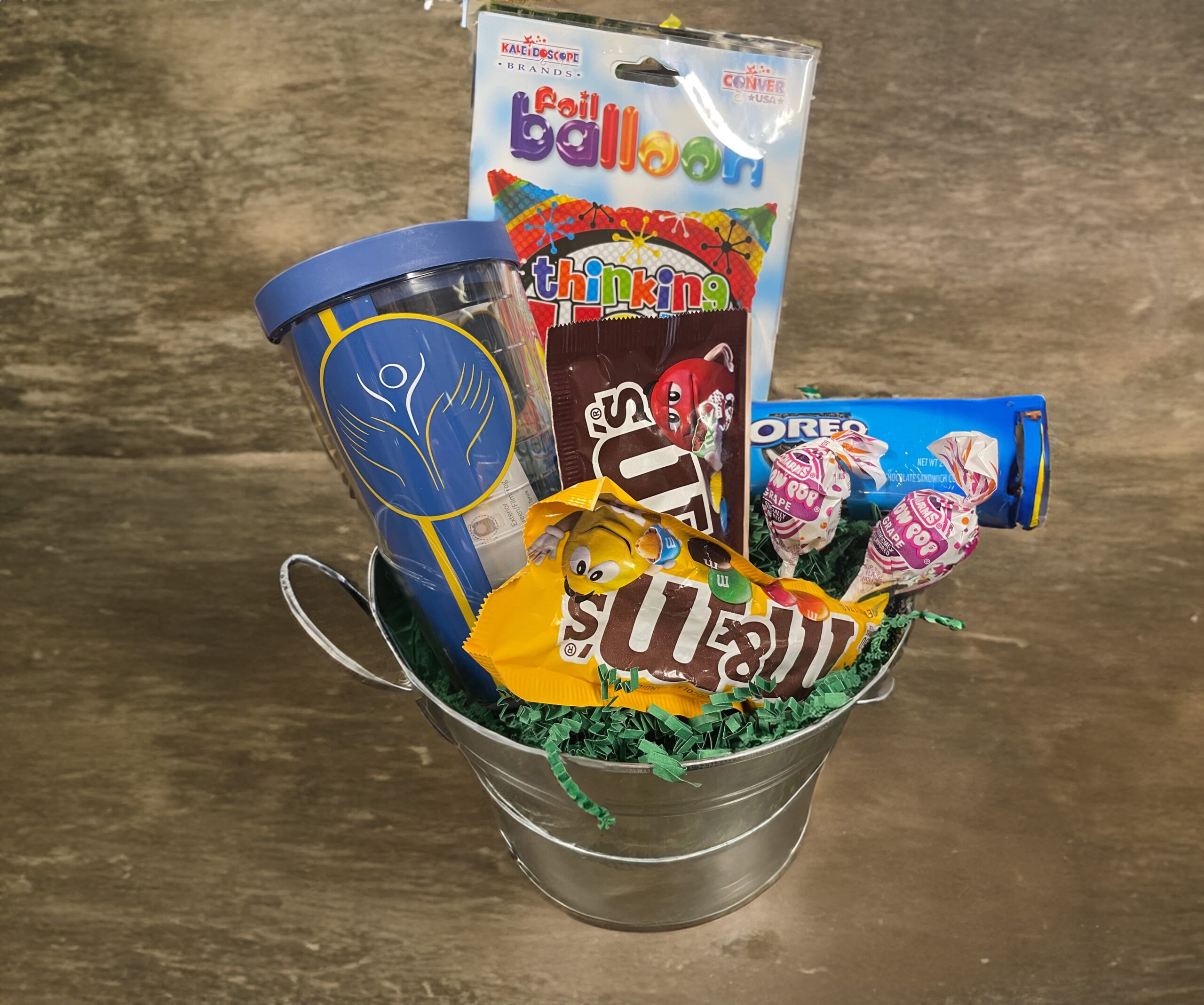 A small metal bucket filled with a variety of items, including a blue travel mug, a packet of foil balloons, a pack of trail mix, an Oreos packet, a yellow pack of peanut M&M's, and two lollipops, set against a wooden background.