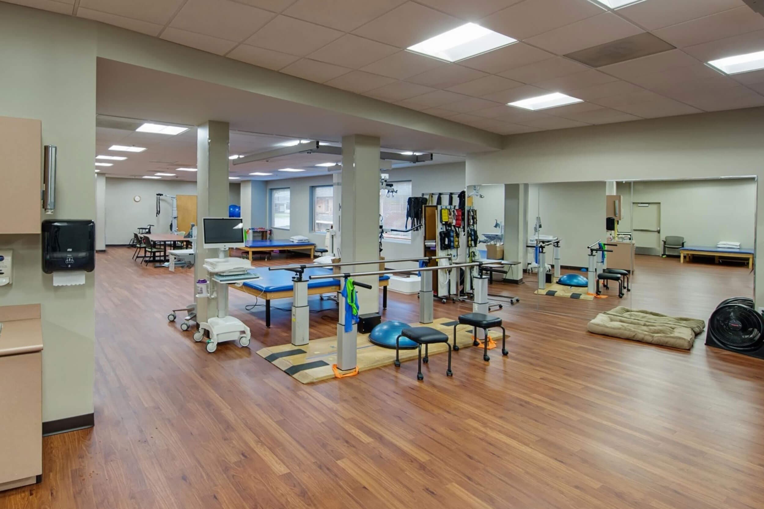 A spacious physical therapy gym with wooden floors, various exercise equipment including parallel bars, mats, and weights. The room is well-lit with large mirrors and windows, creating an open and welcoming environment.