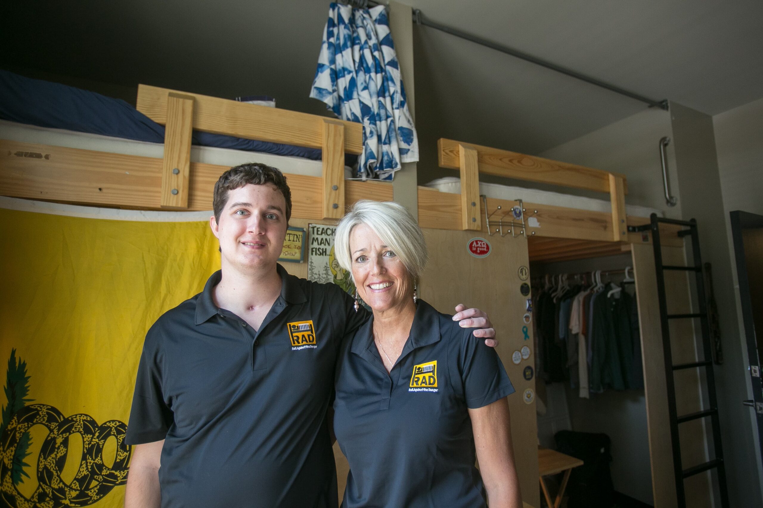 Two people wearing black shirts with yellow logos stand smiling in a dorm room. The room has a lofted bed, a wooden closet, and a yellow flag hanging on the wall. Assorted clothing and a small table are visible in the background.