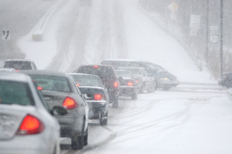 Cars drive in a line on a snow-covered road during a snowstorm, with reduced visibility and overcast skies.