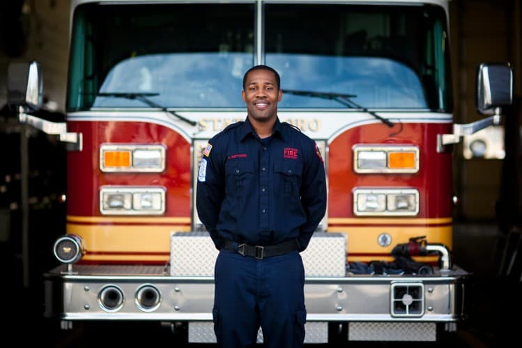 A firefighter in uniform stands smiling in front of a red fire truck inside a garage. The truck has large headlights and a chrome bumper. The firefighter has short hair and is standing with hands clasped.