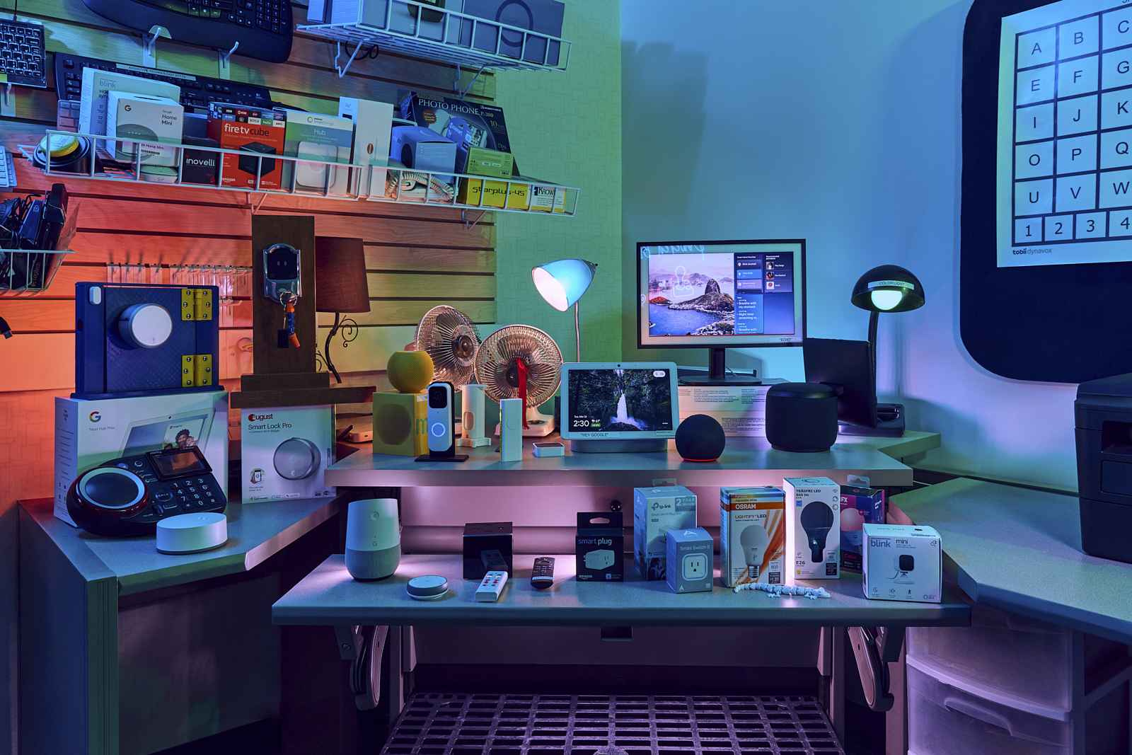 A tech store display featuring various smart home devices. Items include speakers, cameras, and other gadgets on a desk, with a computer monitor and shelves holding more tech products against the wall. The lighting is a mix of warm and cool tones.