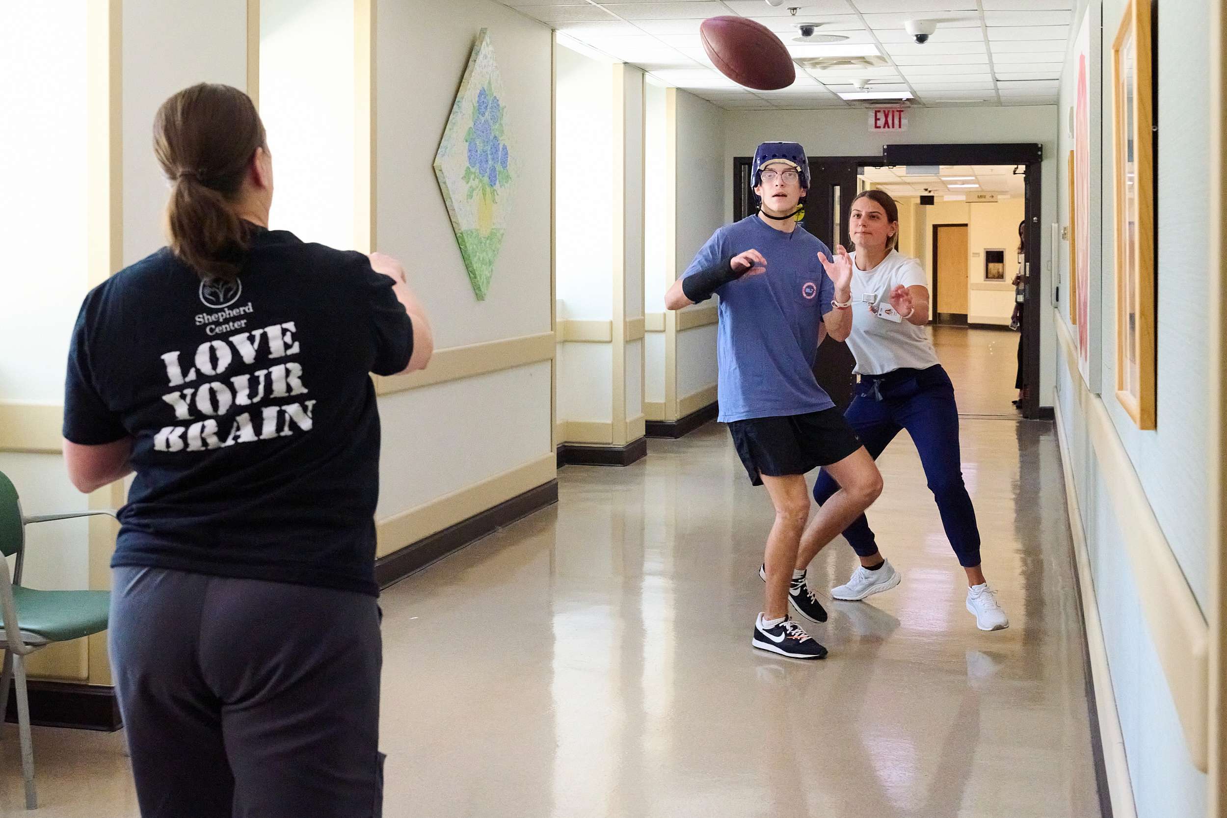 Three people are in a hallway, involved in throwing and catching a football. Two are on one side, with one wearing a helmet and another in a casual shirt. The third person, wearing a shirt with 