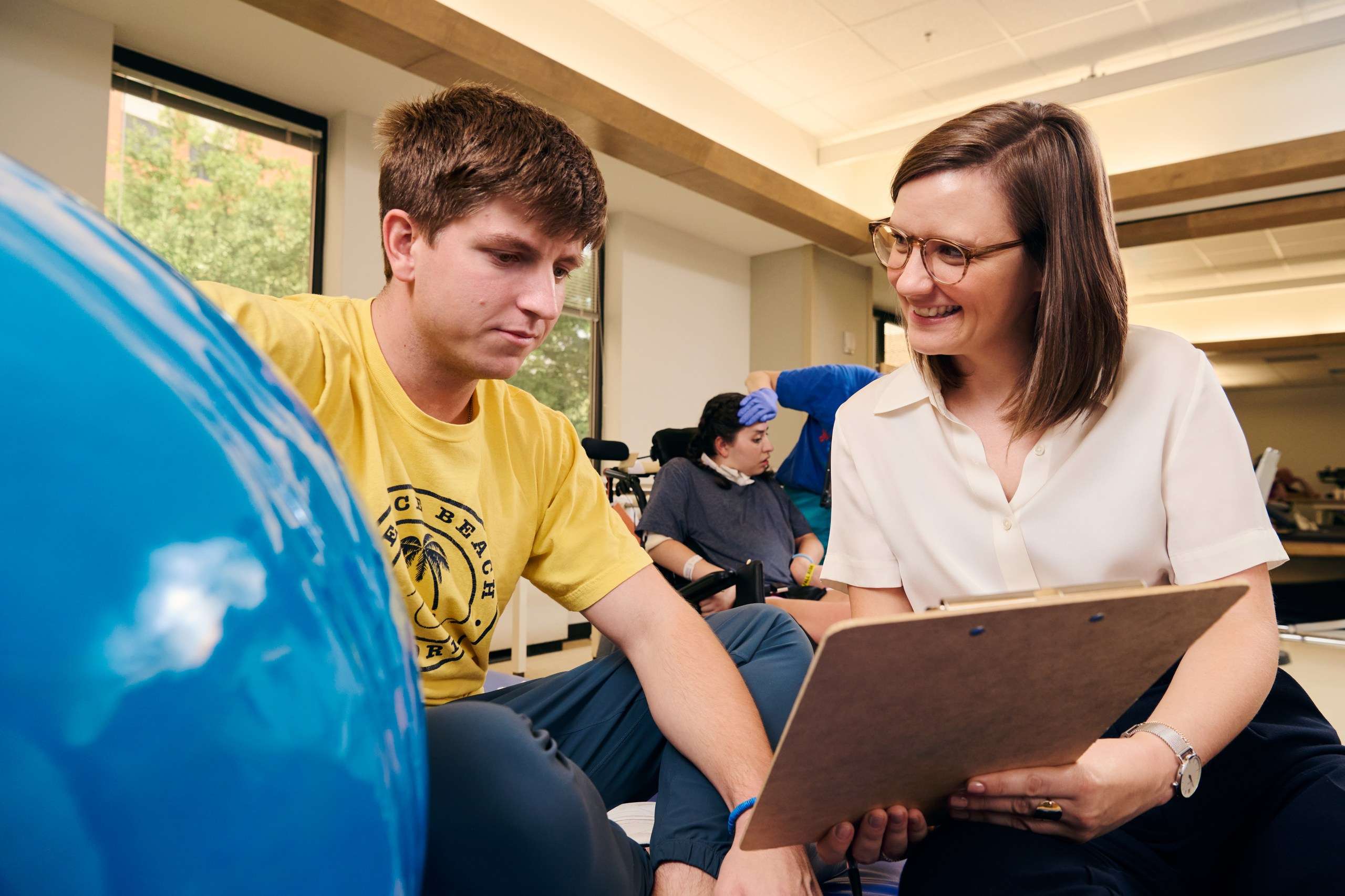 A woman holding a clipboard smiles at a man in a yellow shirt next to a large blue exercise ball. In the background, another person in a blue shirt is seated, visibly focused on a task. The setting appears to be a therapy or rehabilitation room.