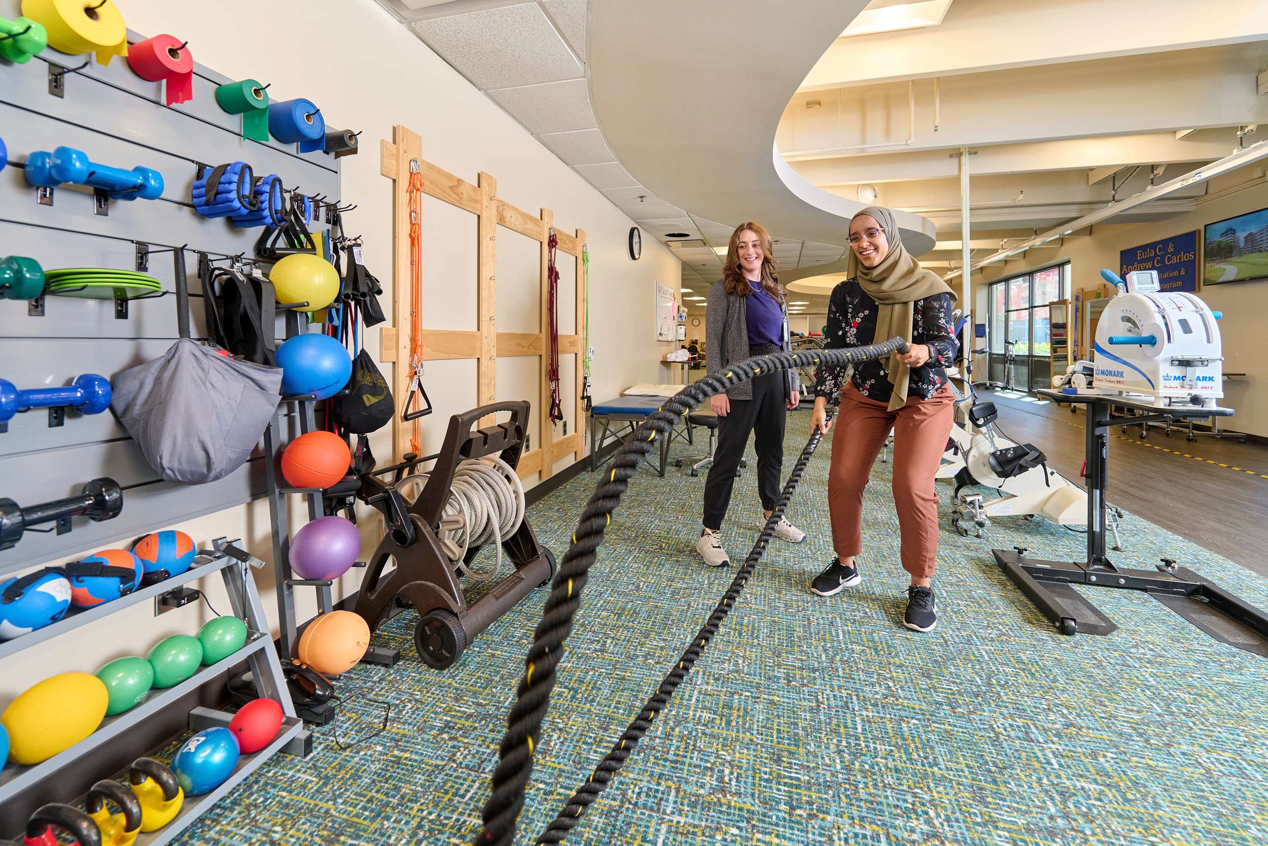 Two women are in a gym, where one uses battle ropes while the other supervises. The room is equipped with various fitness equipment, including weights, balls, and exercise machines. The atmosphere is energetic and focused.