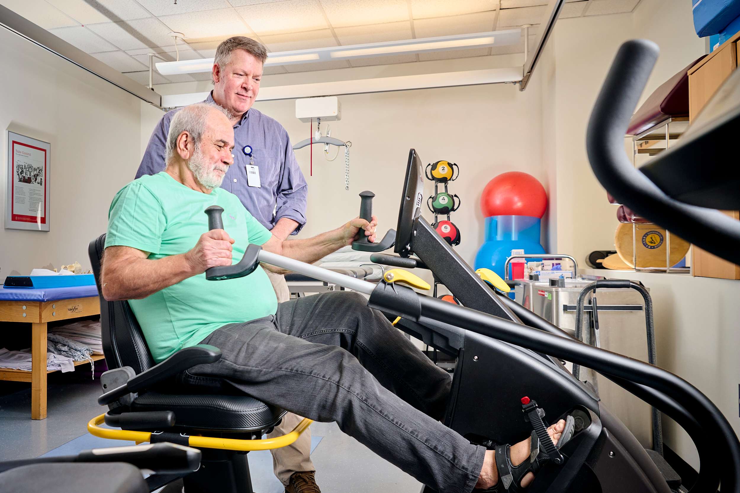 An older man in a green shirt uses exercise equipment in a fitness area, assisted by a man in a blue shirt. Behind them are stacked colorful medicine balls and gym mats. The setting is well-lit and equipped for physical activity.