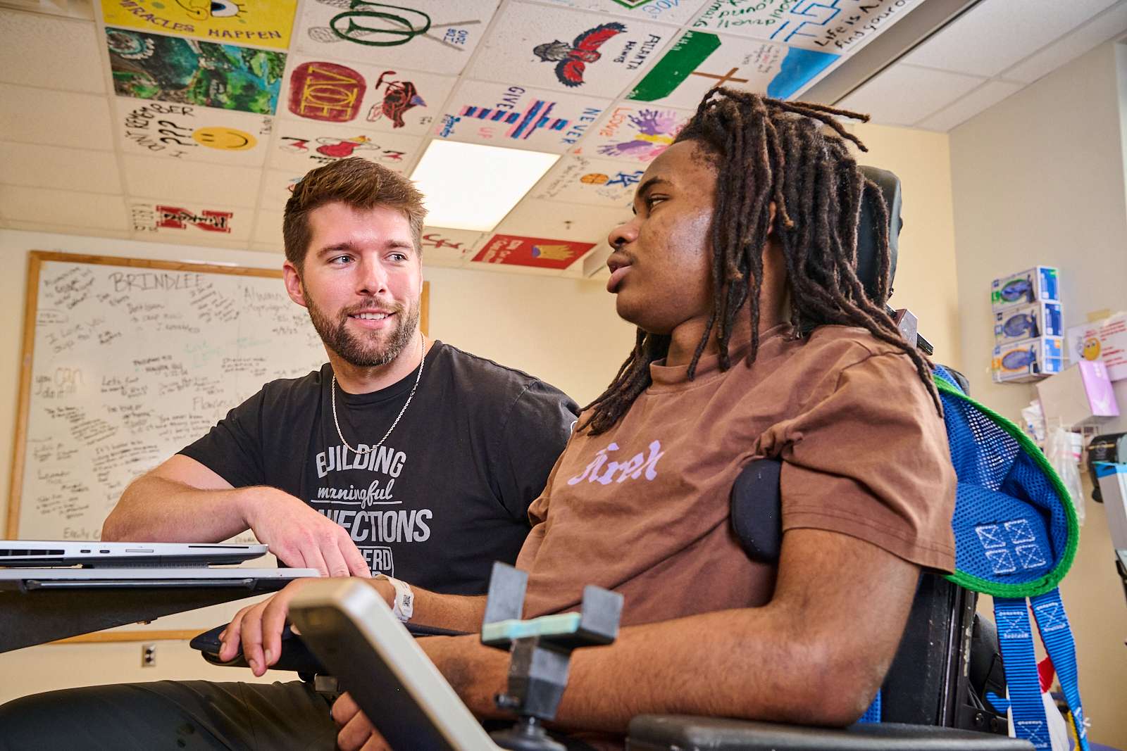 Two men are engaged in conversation at a desk. One man with a beard and a T-shirt is leaning towards the other, who has long hair and is seated in a wheelchair. They are in a room with a whiteboard and ceiling tiles decorated with colorful art.