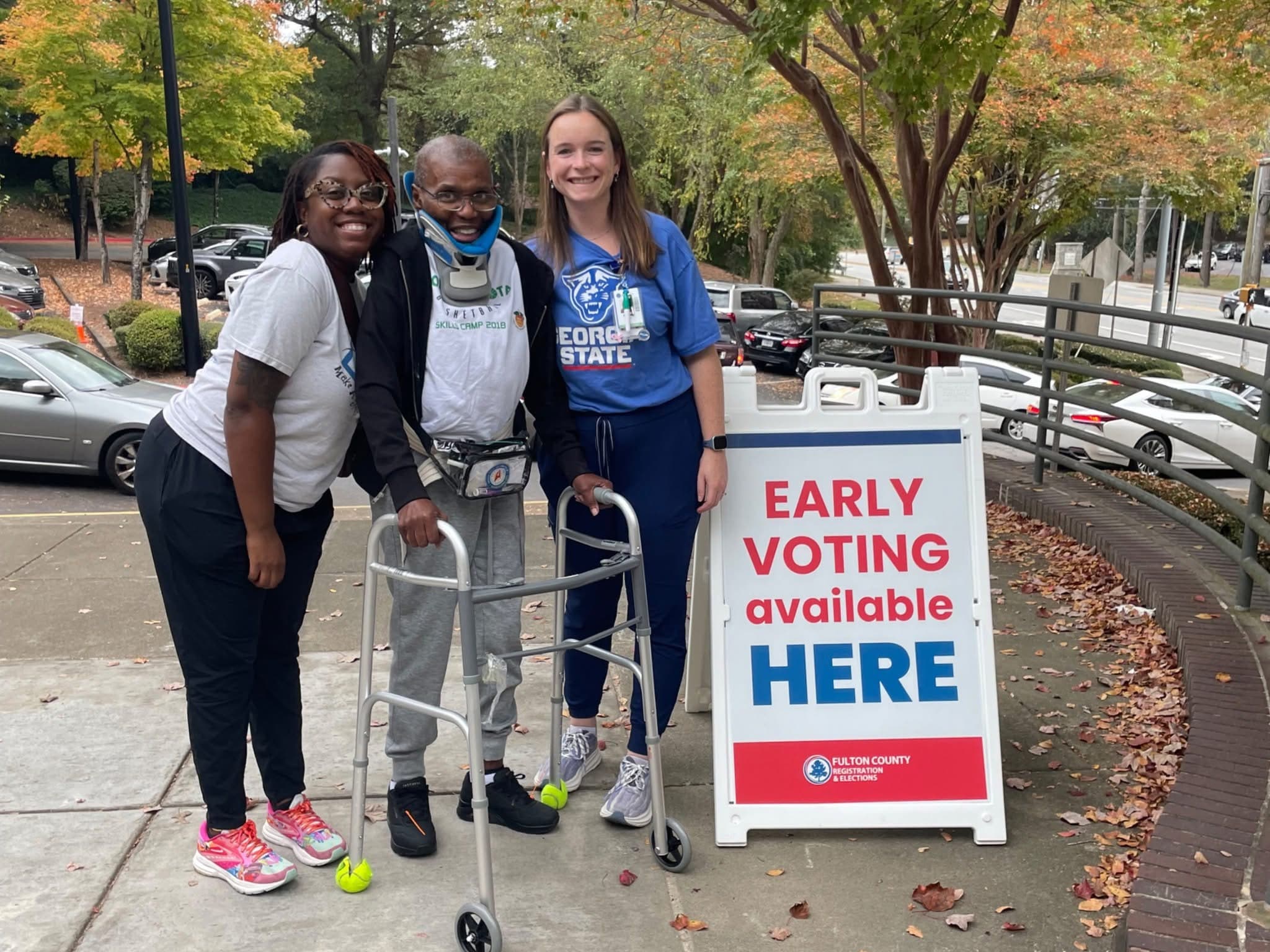 Three individuals stand beside a person with a walker in front of a sign that reads 