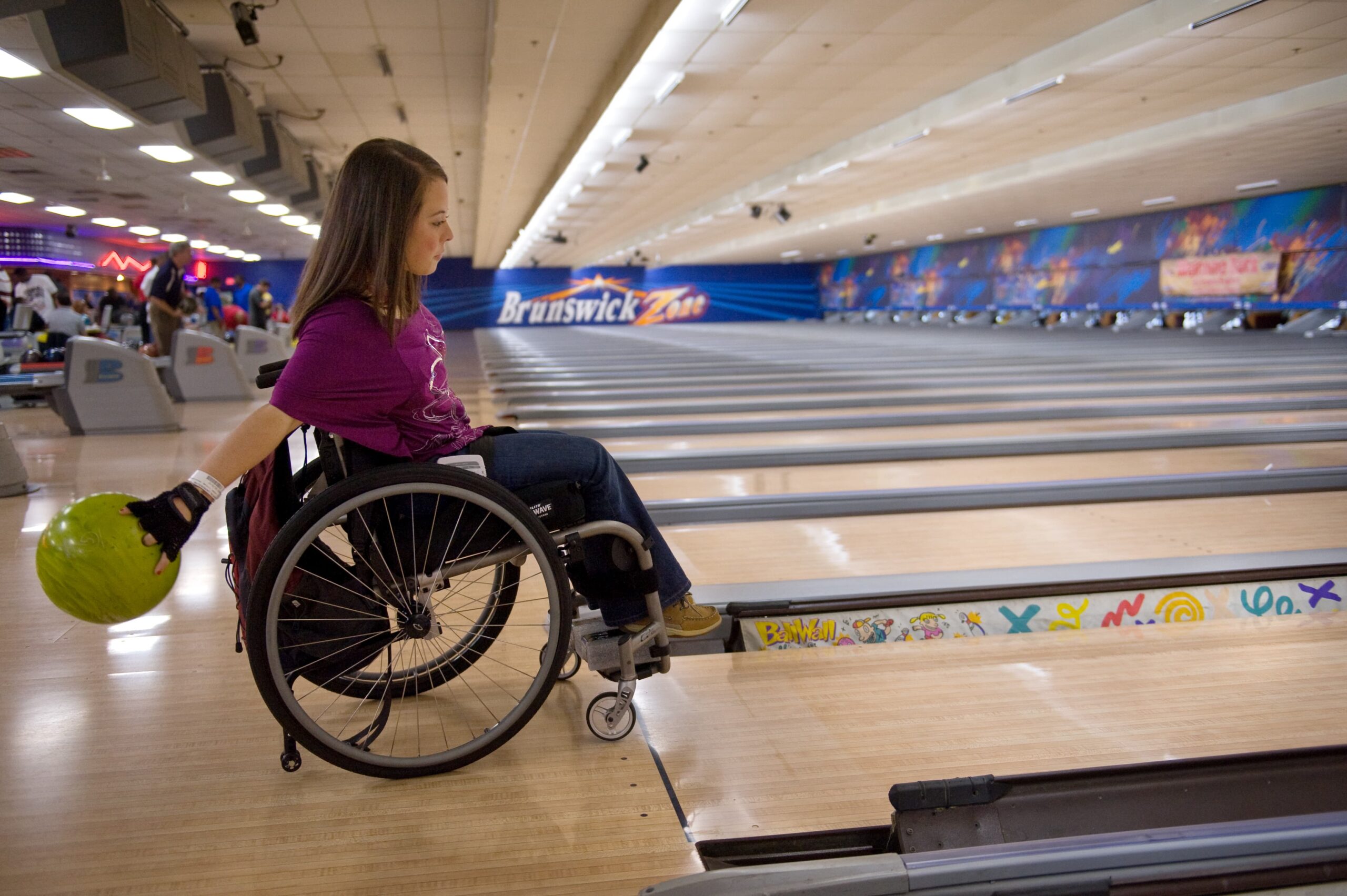 A woman in a wheelchair is at a bowling alley, holding a green bowling ball. She is positioned near the lane, preparing to bowl. The alley is brightly lit, with colorful designs on the side and more lanes in the background.