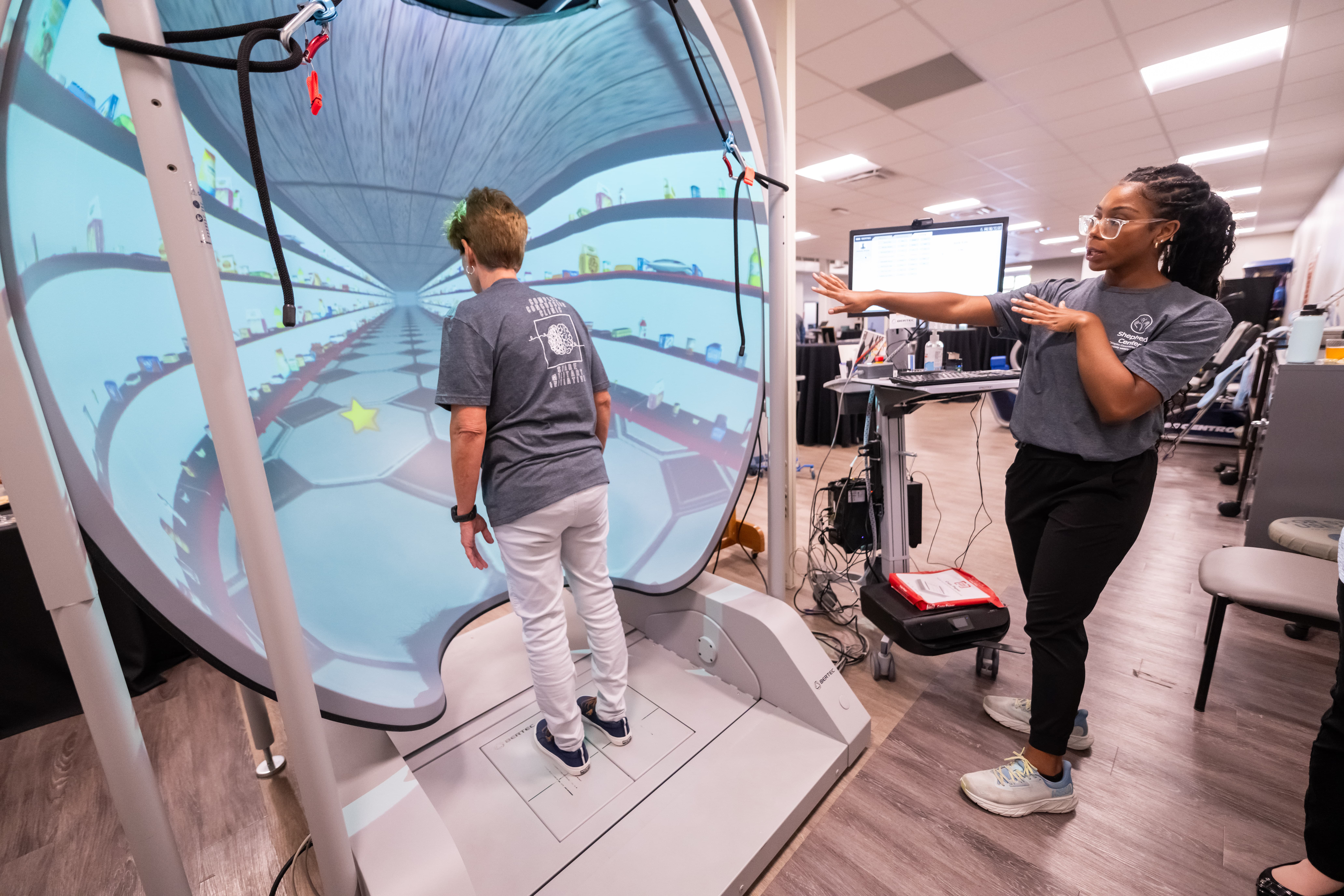 A person stands inside a large virtual reality dome, facing a projection of a digital environment. Another person nearby gestures towards a screen, providing instructions. Both wear casual clothing and appear to be engaged in a tech demonstration.