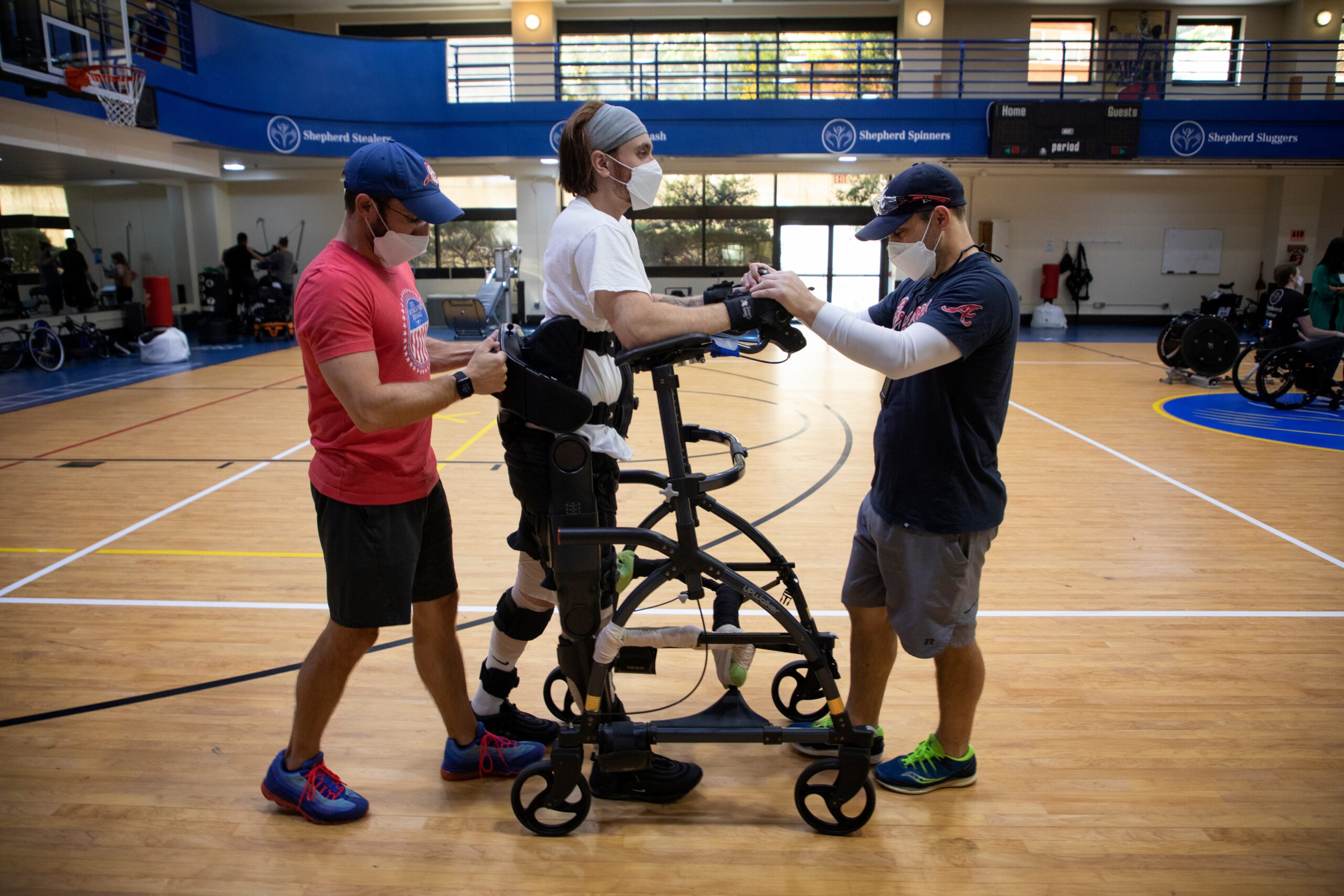 Three people are on a basketball court in a gym. Two of them are assisting a person with mobility aids to stand. All are wearing masks and casual sportswear. Wheelchairs and gym equipment are in the background.