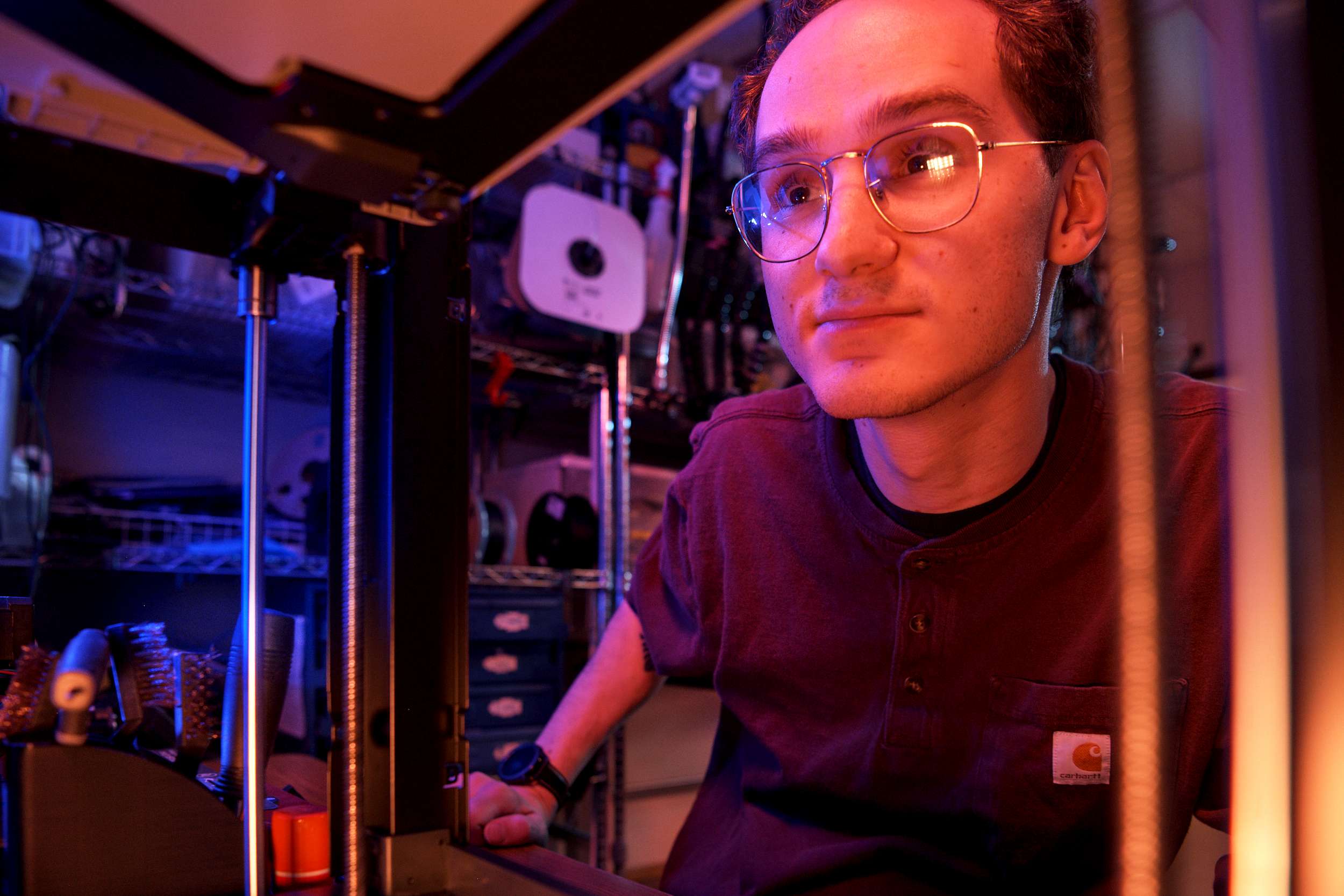 A person with glasses and short hair is leaning close to a 3D printer, appearing focused. The background has shelves with various tools and equipment. The scene is lit with warm, colorful lighting, giving it a technical and creative atmosphere.