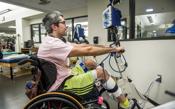 A man in a wheelchair with exercise equipment, using a stationary arm and leg cycle machine in a bright, modern rehabilitation facility. He appears focused, with medical sensors attached to his legs and equipment.