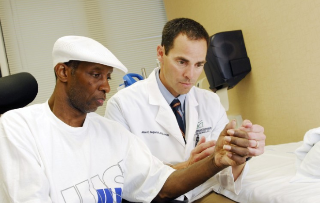 A doctor in a white coat examines a patient's hand in a medical office. The patient is wearing a white cap and T-shirt, while seated beside the doctor, who focuses intently on the patient's hand. A white bed is in the background.