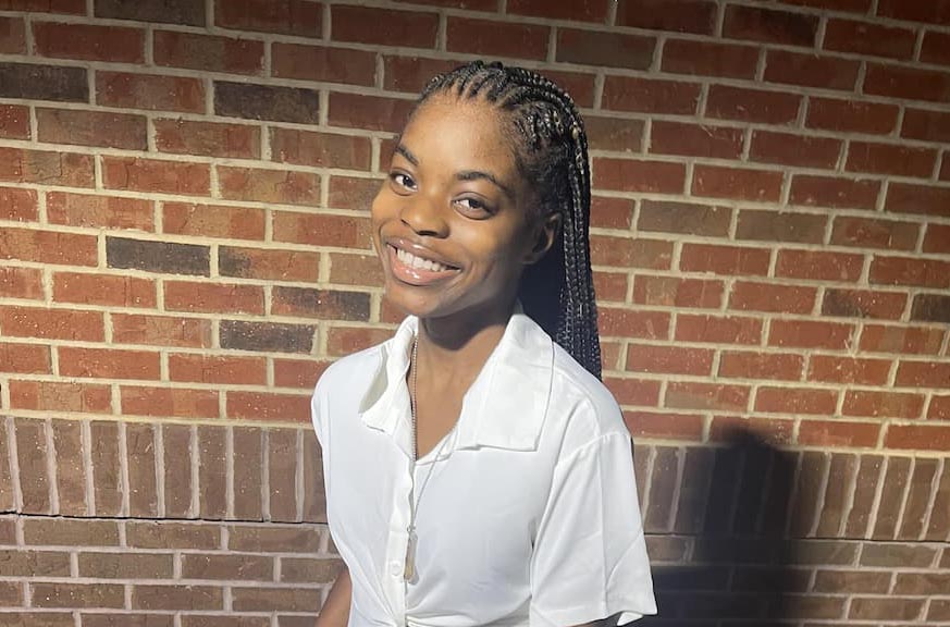 A person with long braided hair smiles in front of a brick wall. They are wearing a white shirt and are beautifully illuminated by sunlight.