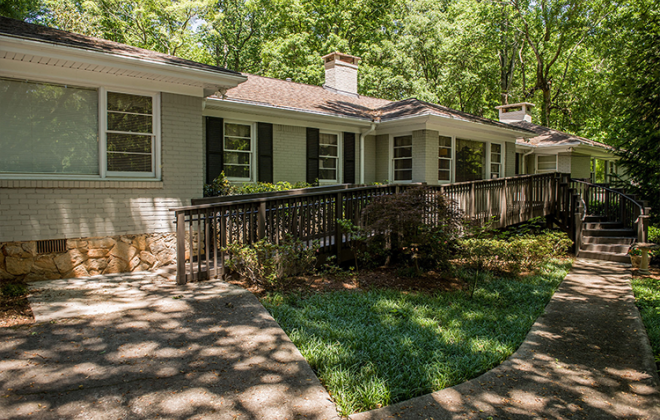 Single-story house with light brick exterior and several large windows, surrounded by greenery and trees. A sloped wooden ramp and stairs lead to the entrance. The setting is serene with dappled sunlight on the paths.