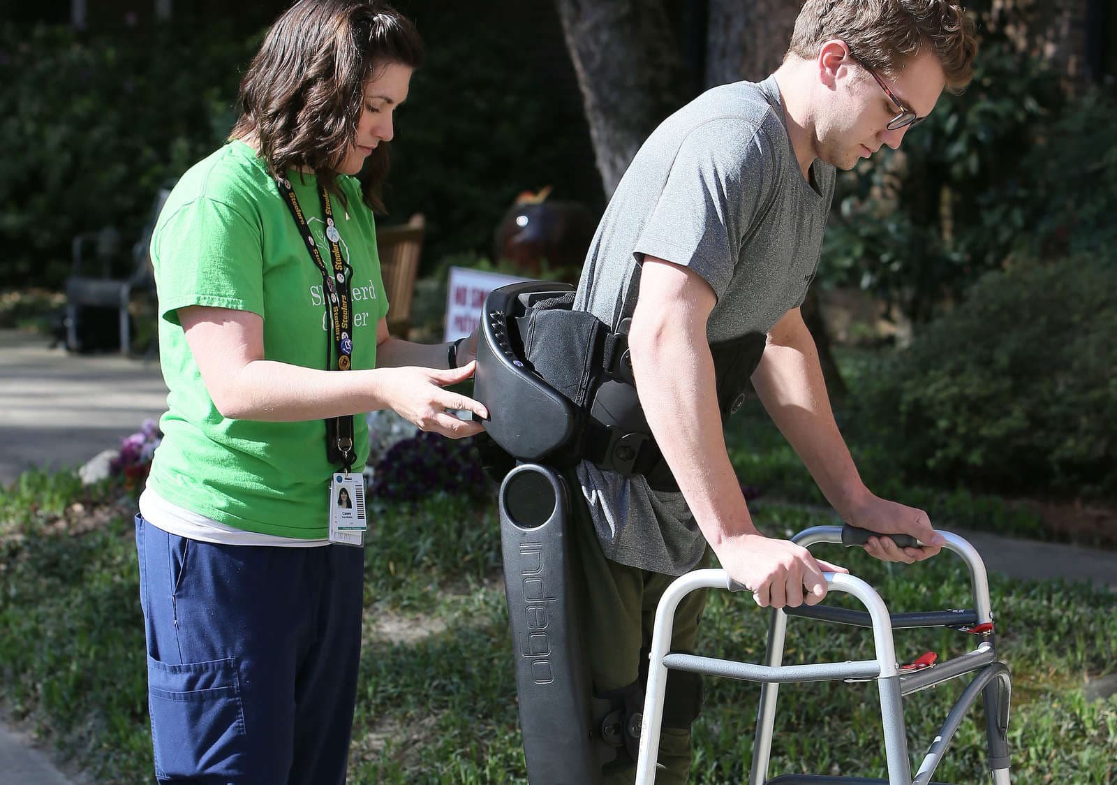 A person assists a man using a walking aid and wearing an exoskeleton device outdoors. The man is holding a walker, and the assistant is wearing a green shirt. They appear to be in a grassy area with trees in the background.
