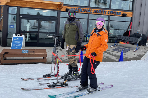Two people in ski gear stand on snow in front of the National Ability Center building. The person on the left wears camouflage pants and a dark jacket, using adaptive ski equipment. The person on the right wears an orange jacket and pink helmet, holding poles.