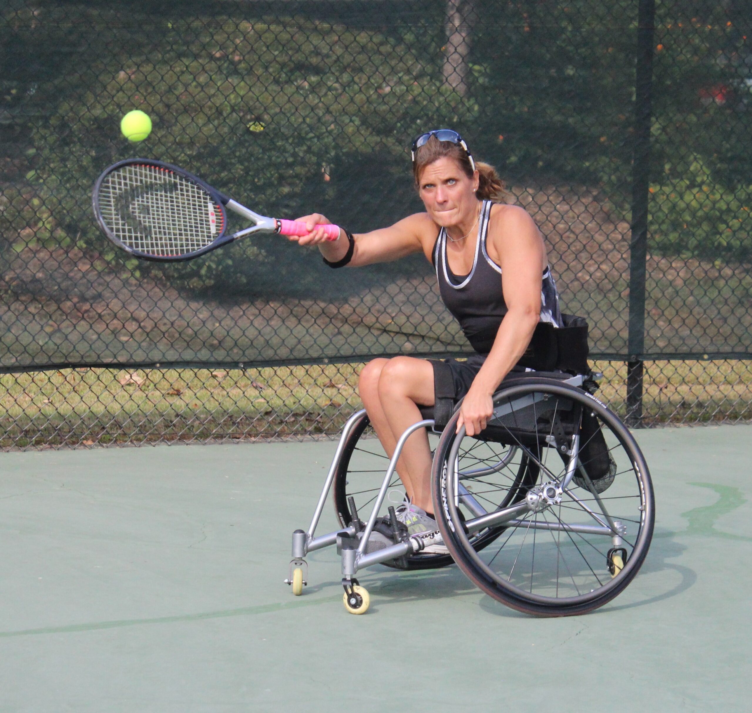 A woman in a wheelchair plays tennis, reaching forward with her racket to hit a tennis ball. She is wearing athletic clothes, sunglasses, and is on an outdoor court with a chain-link fence in the background.
