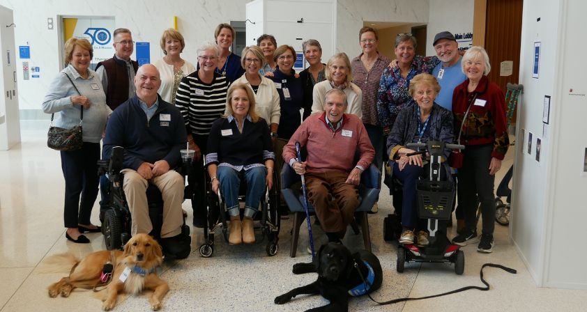 Dr. Apple and former recreation therapists, some in wheelchairs and with service dogs, pose and smile together indoors. They wear nametags and casual clothes, gathered in a bright, modern building.