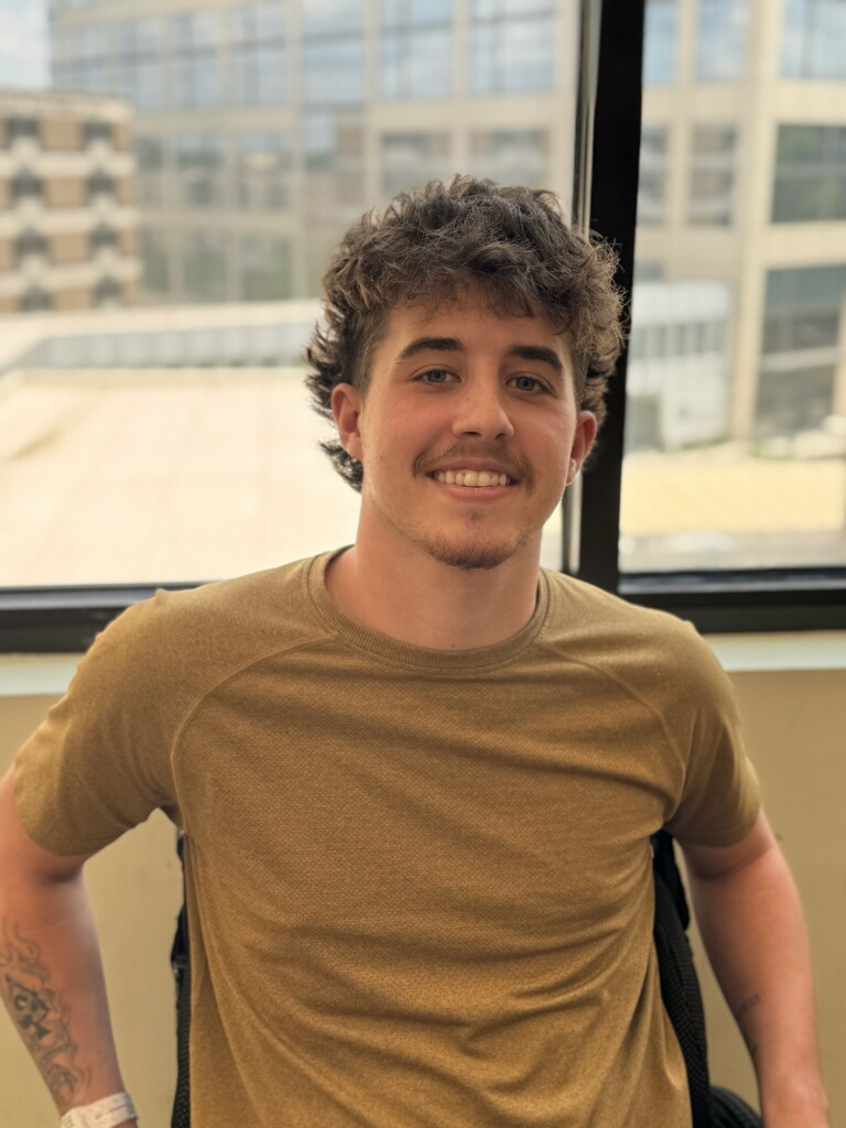 A young man with curly brown hair and a mustache, wearing a light brown t-shirt, smiles while sitting indoors by a window with an urban cityscape in the background.