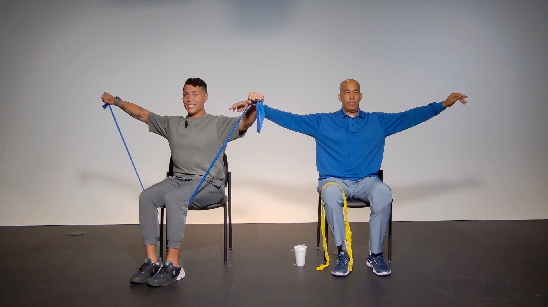 Two people sitting on chairs, each holding resistance bands and extending their arms out to the sides, exercising together in a well-lit room with a plain background. A cup is on the floor beside one chair.