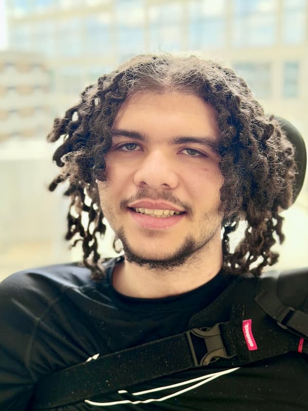 A young man with curly hair smiles at the camera while seated indoors, wearing a black shirt.