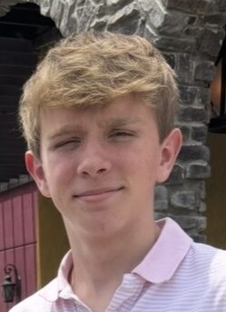 A teenage boy with light brown hair wears a pale pink collared shirt, standing in front of a stone archway, with a slight smile on his face.
