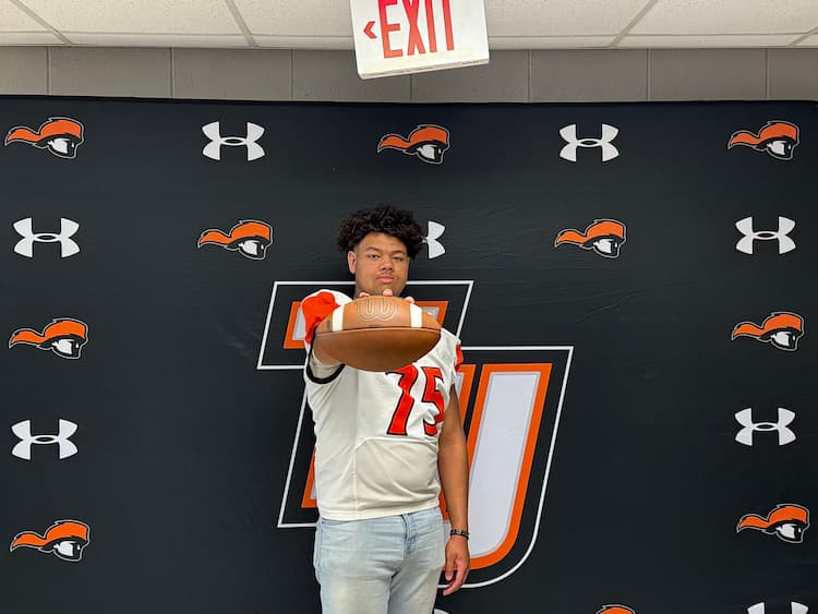 A young man in a football jersey holds a football toward the camera, standing in front of a black backdrop with orange and white logos.