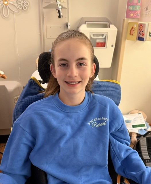 A smiling girl with long brown hair and braces sits in a wheelchair, wearing a blue sweatshirt.