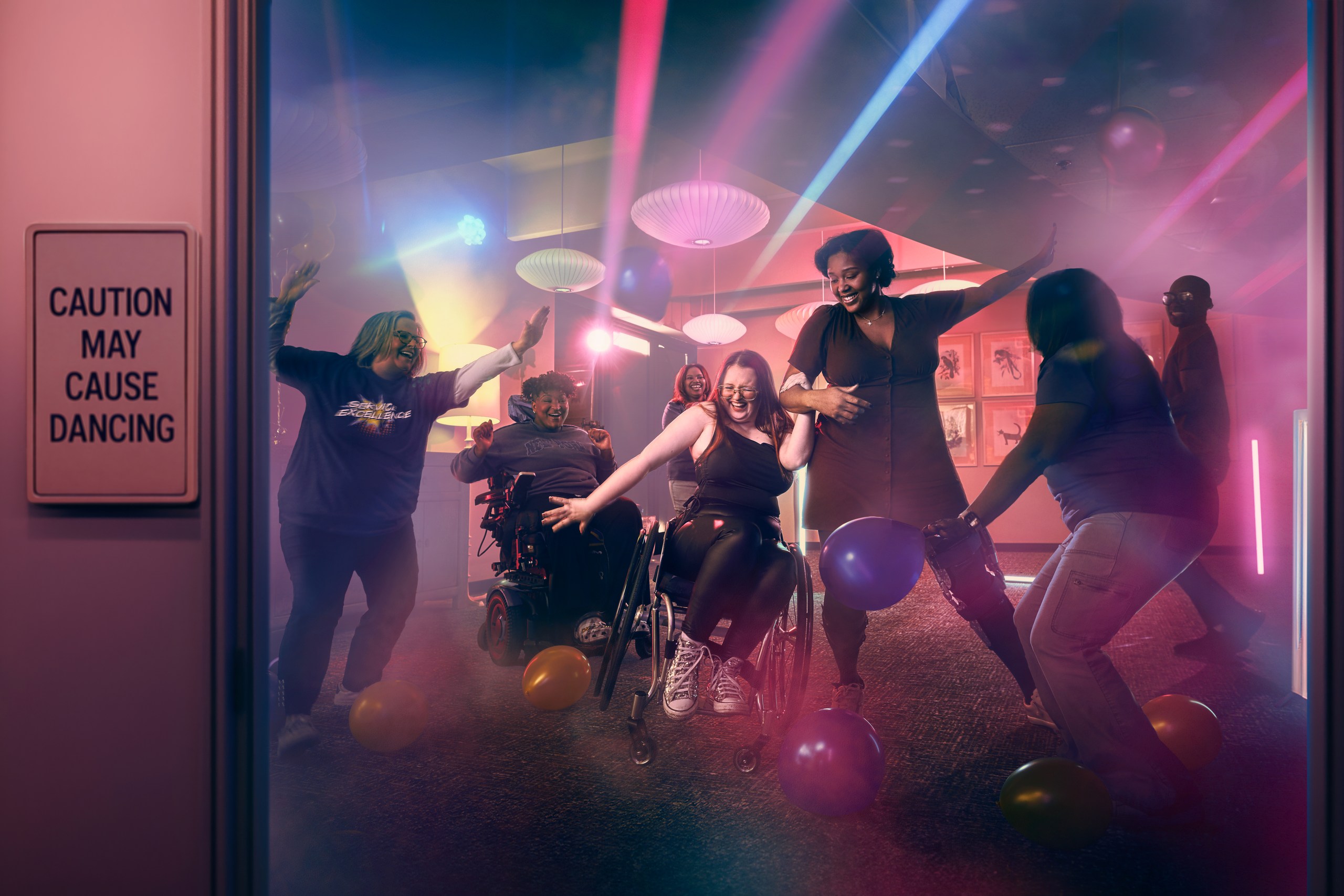 A diverse group of people, including wheelchair users, dance and laugh together in a colorful, neon-lit room with balloons and haze. A sign reads, “Caution may cause dancing,” creating a true sense of belonging for everyone present.