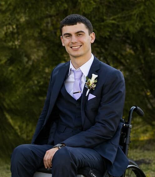 A young man in a suit and tie sits smiling in a wheelchair outdoors, with greenery in the background. He has a boutonniere on his lapel and his hands rest on his lap.