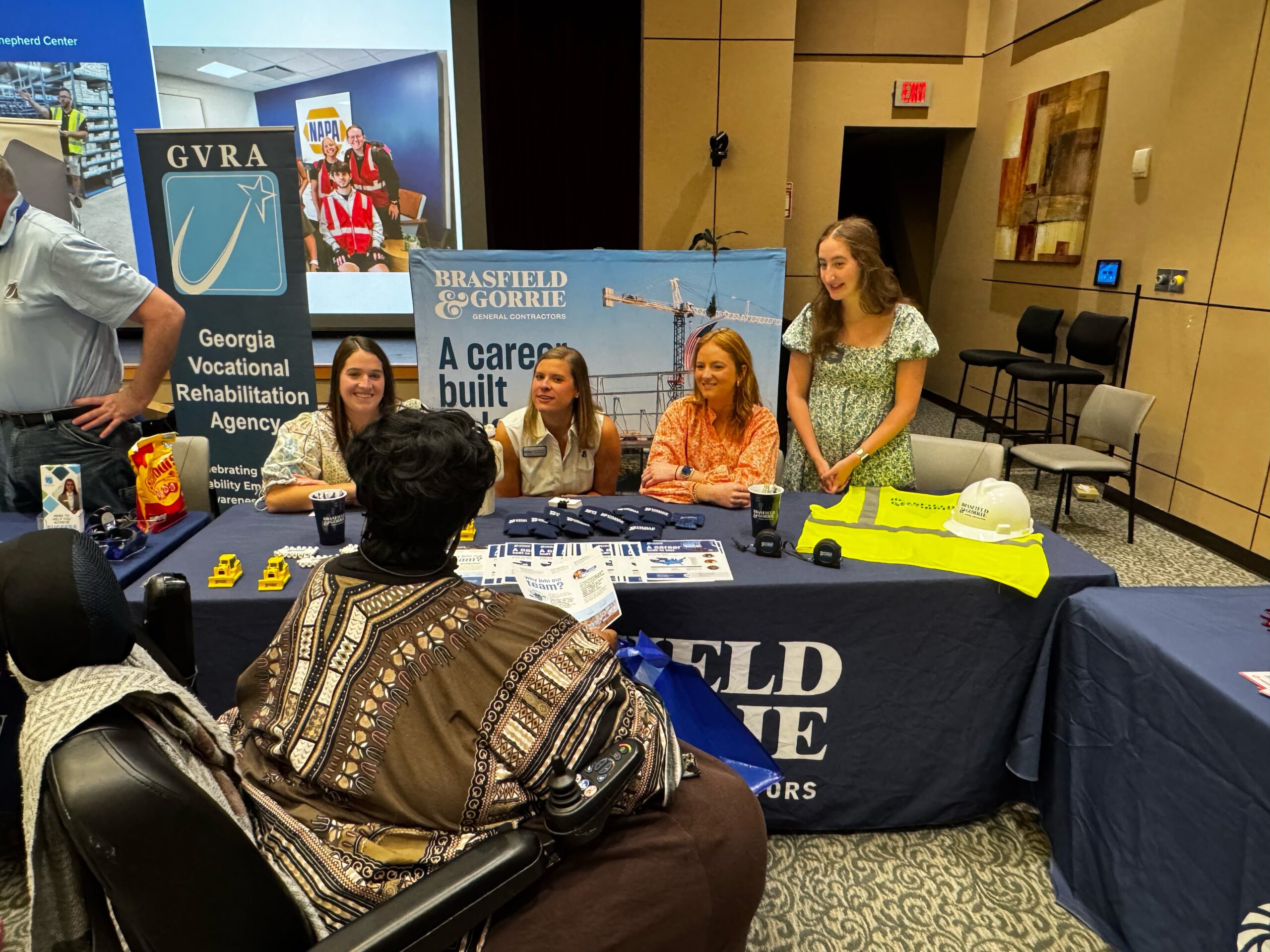 A group of women stand and sit behind a table covered with promotional items and a Brasfield & Gorrie tablecloth at a career fair. A person in a wheelchair is speaking with them. Display banners are visible in the background.
