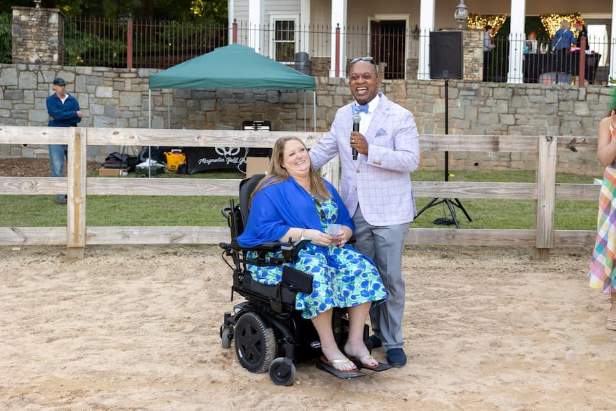 A smiling woman in a blue floral dress sits in a wheelchair beside a man in a light jacket holding a microphone. They are outdoors near a wooden fence, with people and a building in the background.