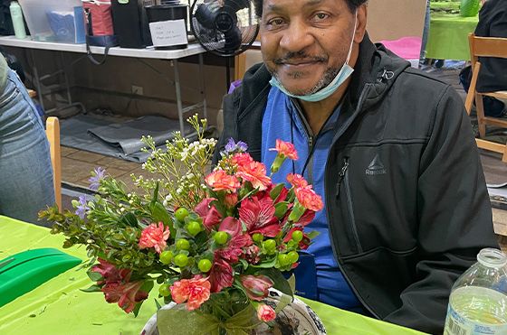 A man sitting at a table with a colorful bouquet of flowers in front of him. He wears a black jacket and blue shirt, and a mask is resting on his chin. The background has various items and a fan.