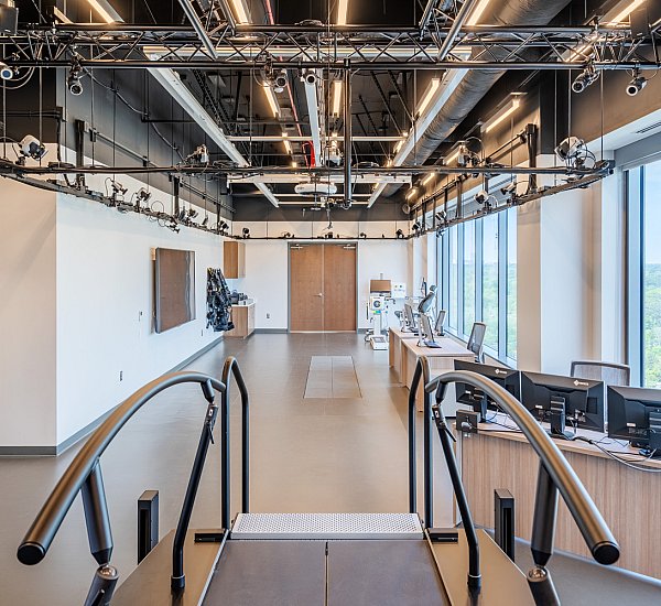 A modern, bright research lab features overhead motion capture cameras, computers on wooden desks, large windows, and a treadmill with handrails at the center. The room is spacious with high ceilings and exposed pipes.