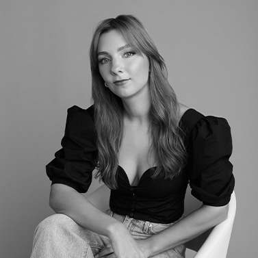 Black and white portrait of a woman with long hair, wearing a puff-sleeve black top and light jeans, sitting on a white chair against a plain background with a relaxed, confident expression.