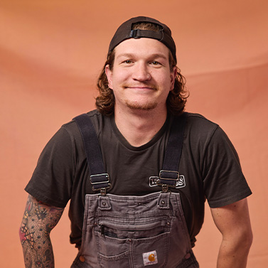 A man with long hair wearing a backward black cap, dark t-shirt, and gray overalls smiles at the camera. He has a tattooed left arm and stands against a peach-colored background.