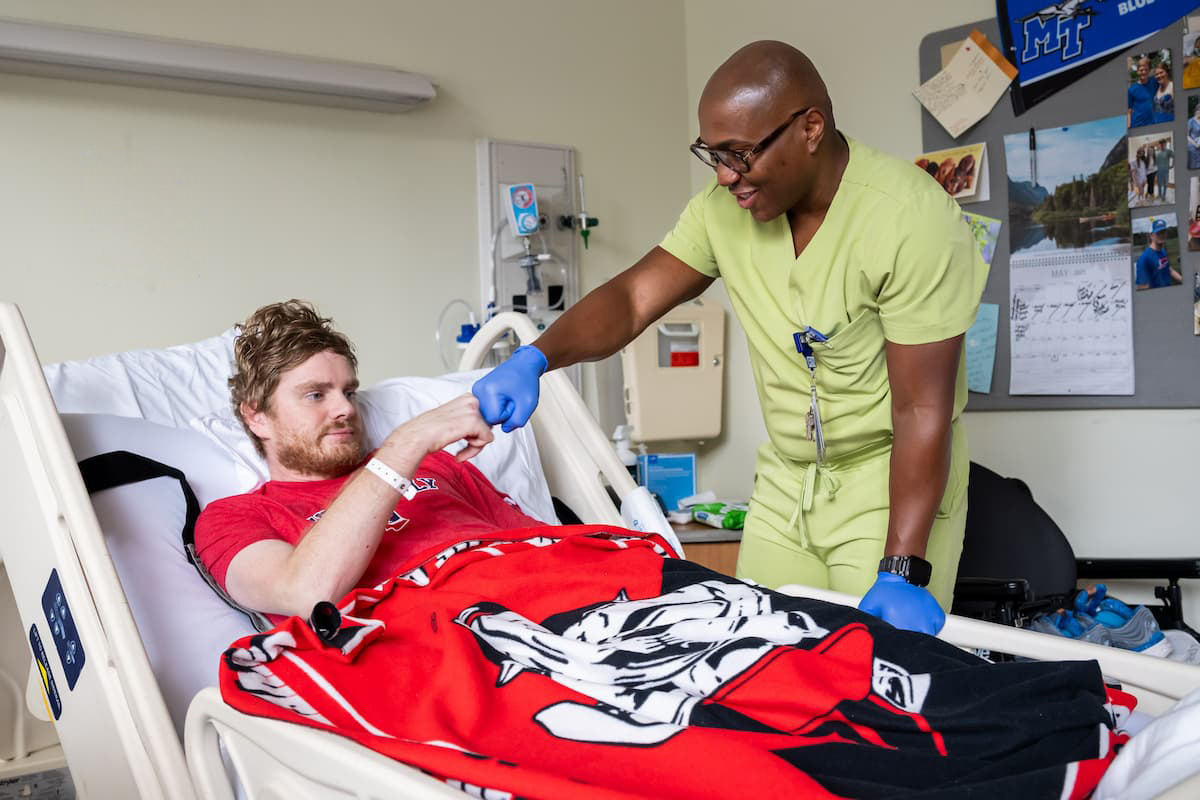 A nurse in green scrubs wearing gloves fist bumps a patient lying in a hospital bed with a red blanket. The patient, wearing a red T-shirt, smiles as they interact in a bright hospital room.