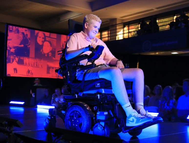 A young person in a motorized wheelchair smiles while spotlighted on a stage, surrounded by blue lighting and an audience. A large screen with faces and images is visible in the background.