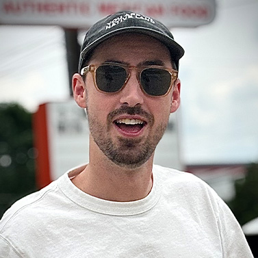A man wearing sunglasses, a black baseball cap, and a white shirt is smiling outdoors. The background is blurred, with some signage and trees visible.