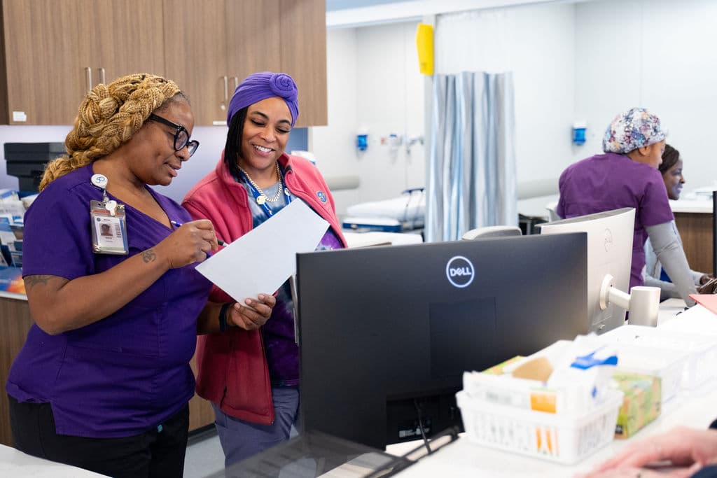 Two healthcare workers in scrubs smile while reviewing a document together at a desk in a medical office. A computer monitor and medical supplies are on the desk, with other staff working in the background.