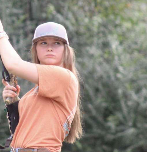 A young person with long hair wearing an orange t-shirt and a light-colored cap is outdoors.