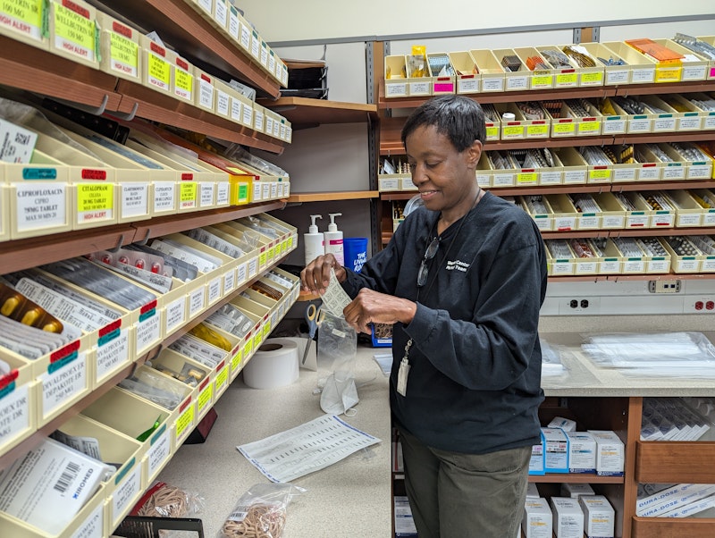 A woman stands in a pharmacy, smiling as she sorts medications into labeled bins. Shelves behind her are filled with various medicine bottles and boxes.
