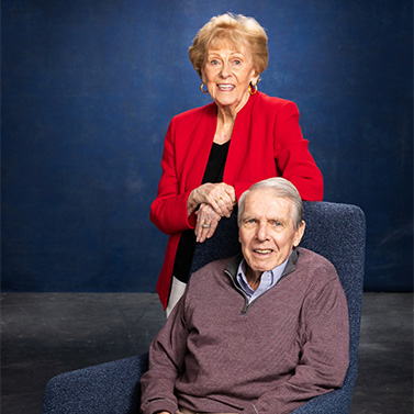 An elderly woman in a red blazer stands smiling behind an elderly man seated in a blue chair, both posing against a dark blue studio backdrop.