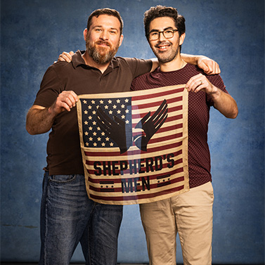 Two men standing side by side, smiling, and holding a small American flag with black wings and the text “SHEPHERD'S MEN” against a blue background.