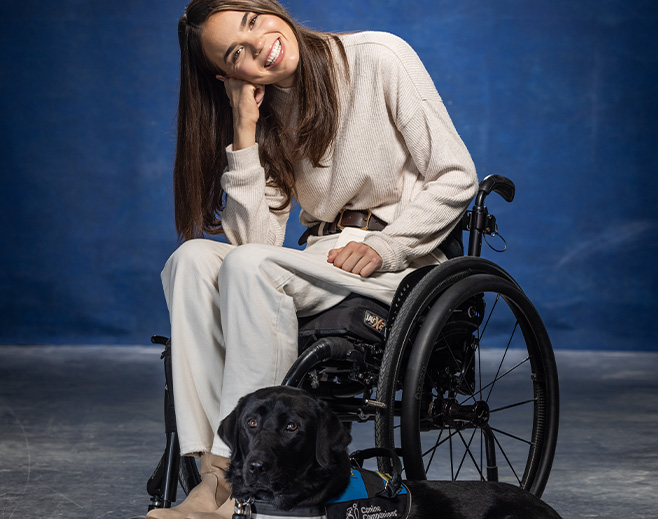 A smiling woman sits in a wheelchair, wearing a light sweater and pants. She leans her head on her hand. A black service dog with a blue vest lies on the floor in front of her. The background is blue.